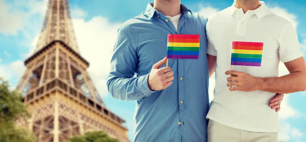 Two male holding a small LGBT flag while in background is the Eiffel Tower. 