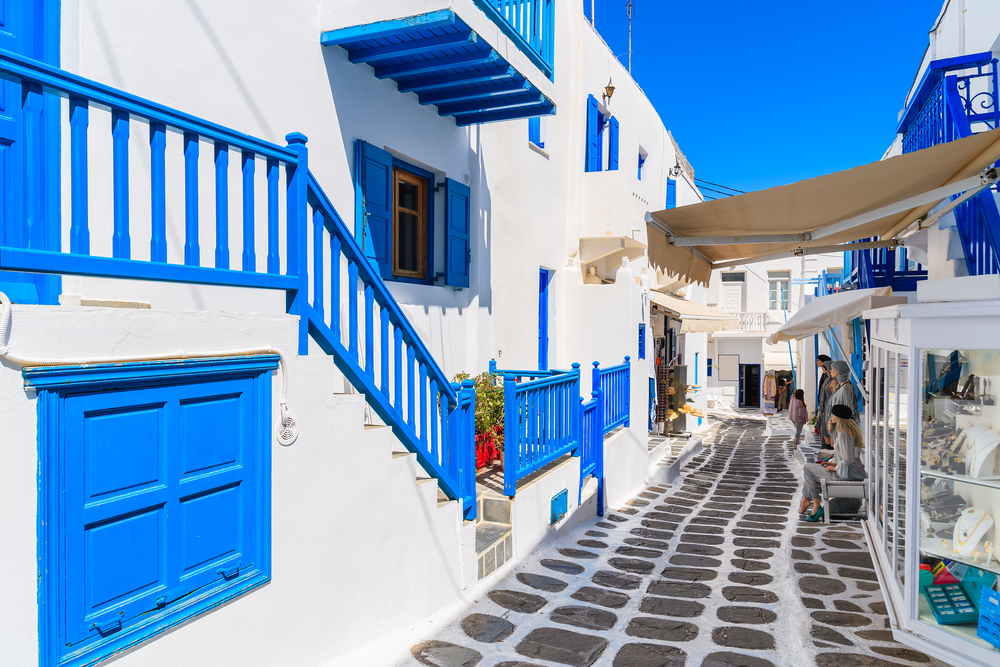 Empty street of Mykonos with its white buildings and blue doors and a bright sky overhead, seen during the least busy time to visit Greece