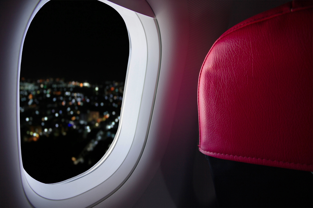 Airplane passenger window with red seat headrest showing as lights from the city below at night peek through the opening for a guide on red eye flights