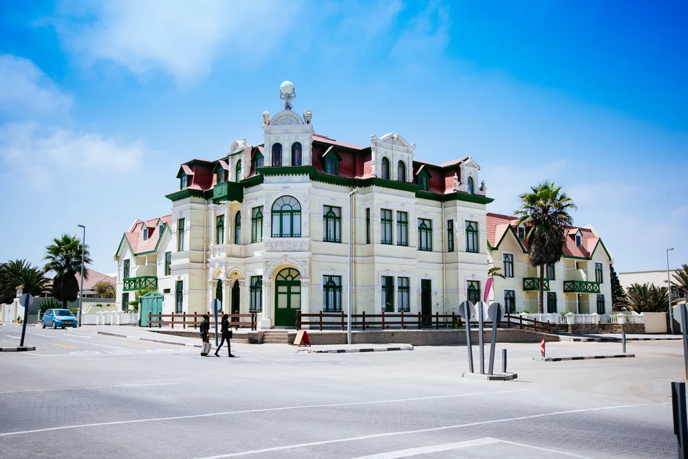 two persons walking on the side of a white old building in Swakopmund, one of the best areas to stay in Namibia, a human figure carrying a globe is at the top of the building.