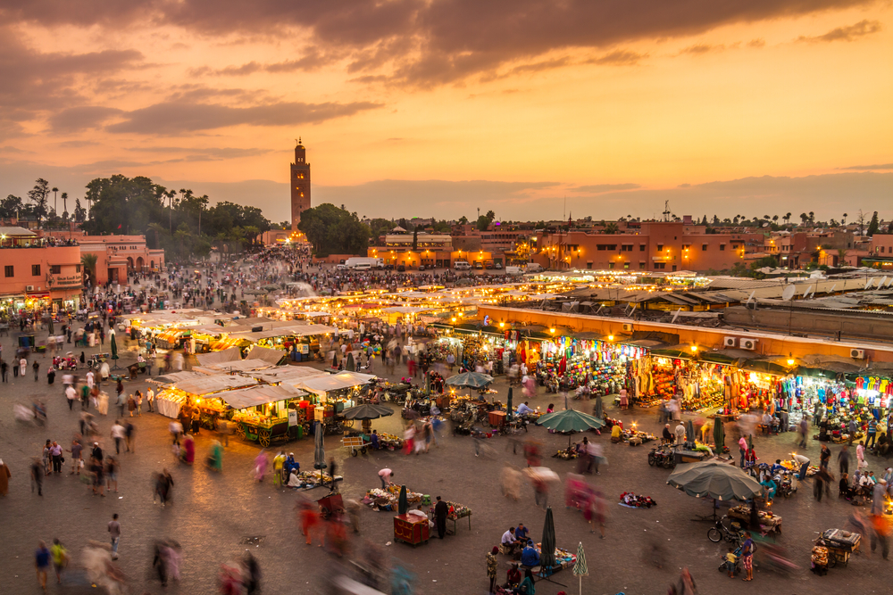 A market area during sunset in Medina, of the best areas to stay in Marrakech, people are busy selling and buying and the lights from store stalls illuminates the area.