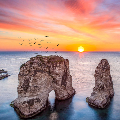 a huge rock formation peaking out of the sea where birds can be seen hovering above it, during a sunset of the best time to visit Lebanon.
