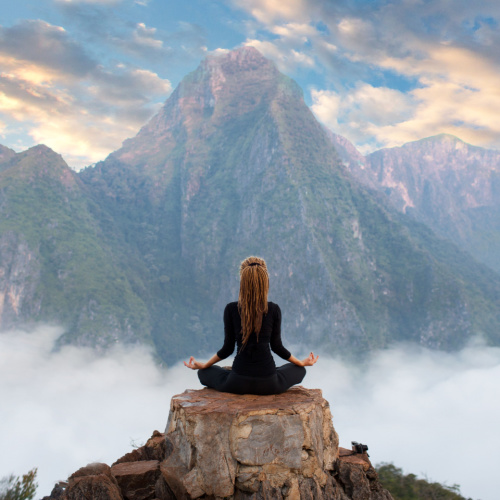 a woman performing yoga while on a rock on top of a mountain where in the other side is the peak of another mountain above the clouds, scenery during the best time to visit Laos.