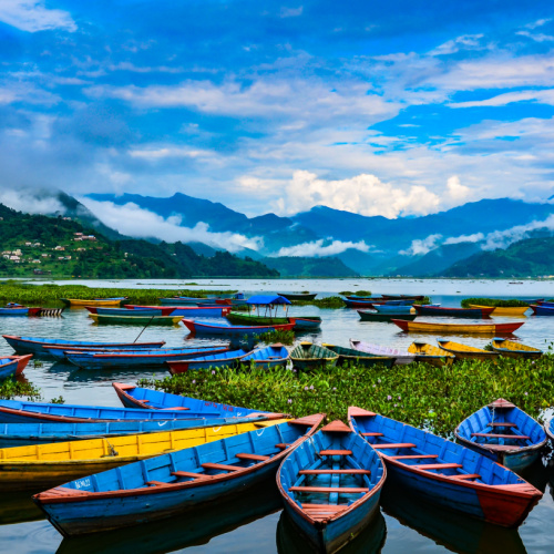 small fishing boats docked on the shallow area of a lake where tall cloudy mountains can be seen in background during the best time to visit Nepal.