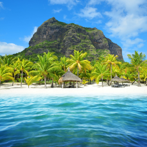 native huts on the beach with white sand, clear water, and palm trees, and a mountain can be seen in background, during the best time to visit Mauritius.