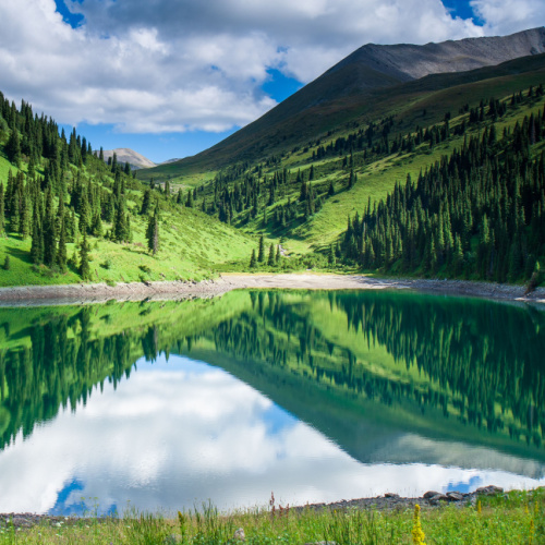 a beautiful scenery during the best time to visit Kazakhstan, where a lake is reflecting green mountains with pine trees.