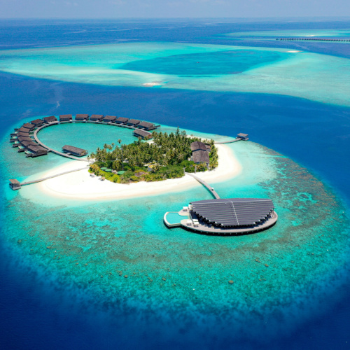 a small island that looks like a paradise with accommodations arranged in an arch while a small area at the center has palm trees, seen during a hot afternoon in the best time to visit Maldives.