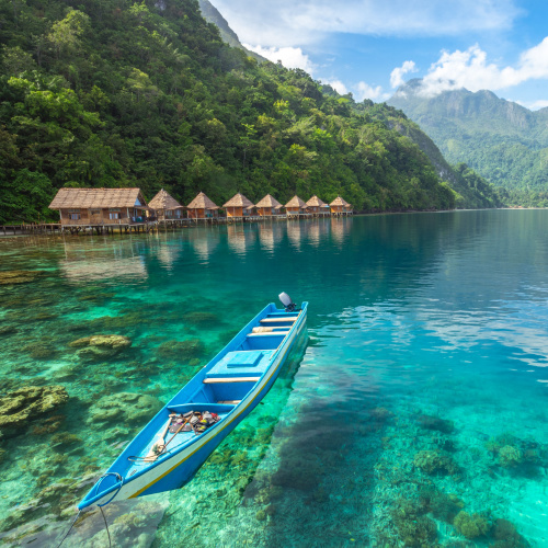 a small blue boat floating on a crystal clear water during the best time to visit Indonesia, where in background are native huts and mountains.