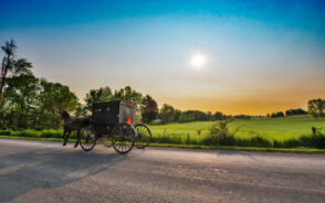 For a guide titled Best Time to Visit Amish Country, PA, a black horse-drawn buggy going down the road on a clear summer evening