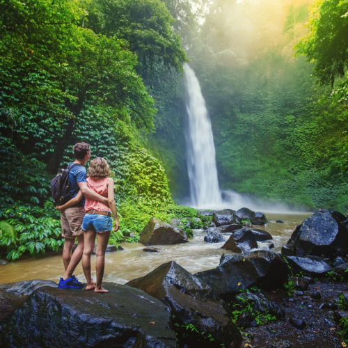 a couple standing on a rock in the river while looking at the falls, surrounded by greenery, during the best time to visit Malaysia.