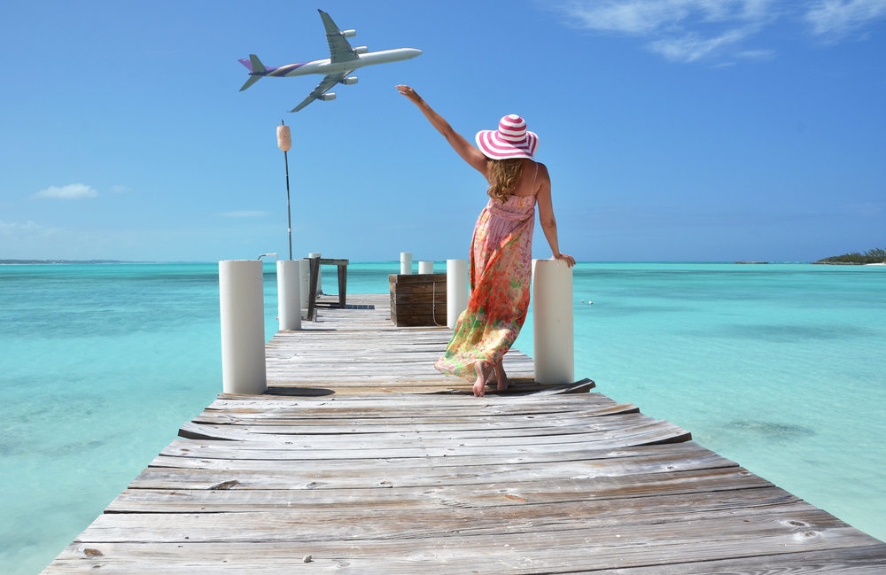 A woman wearing a beach dress while standing on a wooden dock, waving her hand on a passing airplane above, a concept image for a guide about how long is the flight to the Bahamas.