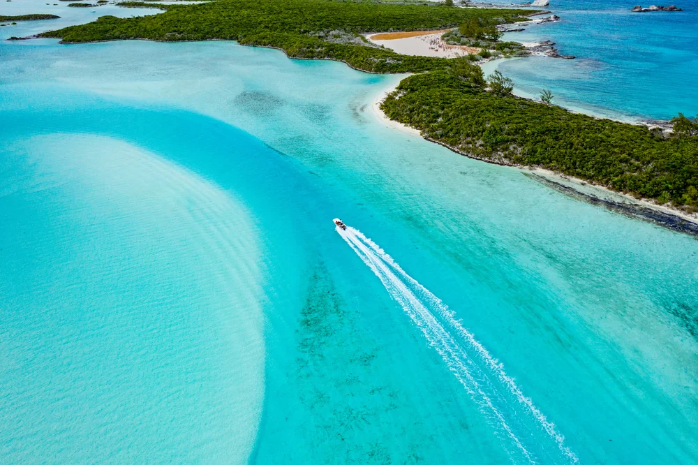 Aerial photo of an island with a crystal clear water and white sand, where a speedboat can be seen on its waters, an image for a guide about how many are in the Bahamas.