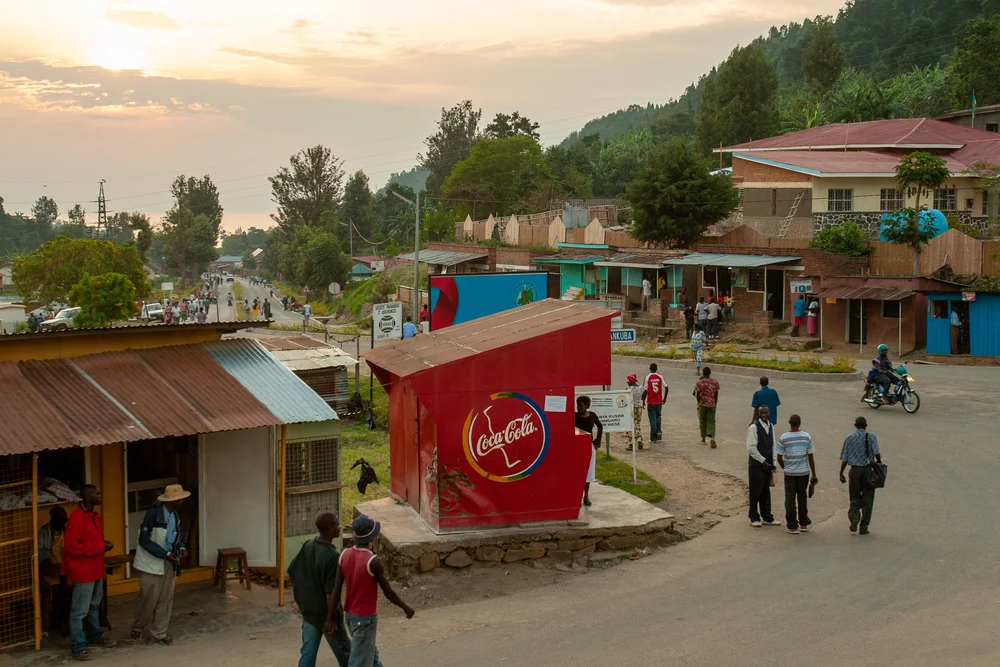 For a piece titled Is Rwanda Safe to Visit, a bunch of villagers walking around a city square on the side of a mountain with little huts surrounding the dirt road