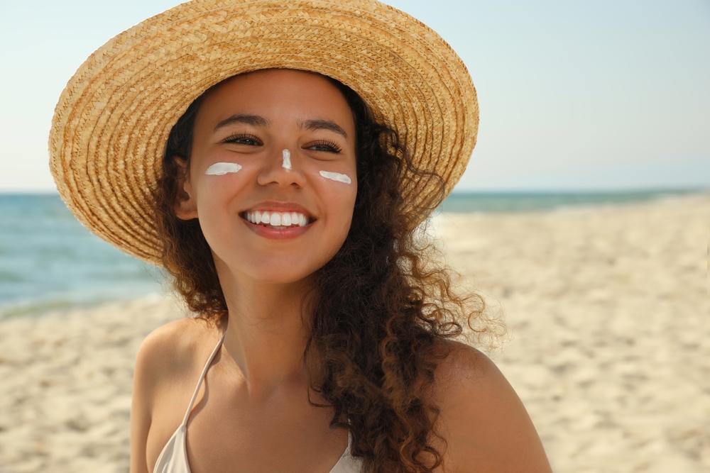 A beautiful woman wearing a hat on the beach, and her face can be seen with sunscreen on her nose and cheeks. 
