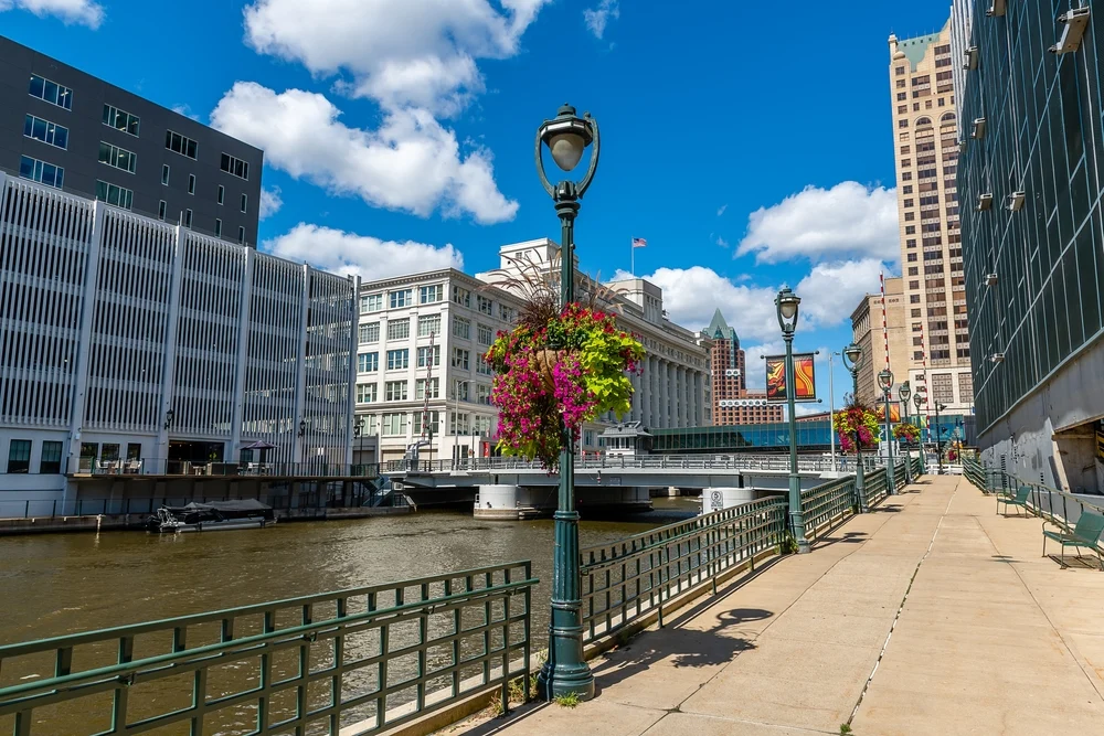 Photo of buildings in the riverwalk district of the city with walkways lining the river and lampposts with flowers hanging from them for a guide titled Is Milwaukee Safe to Visit