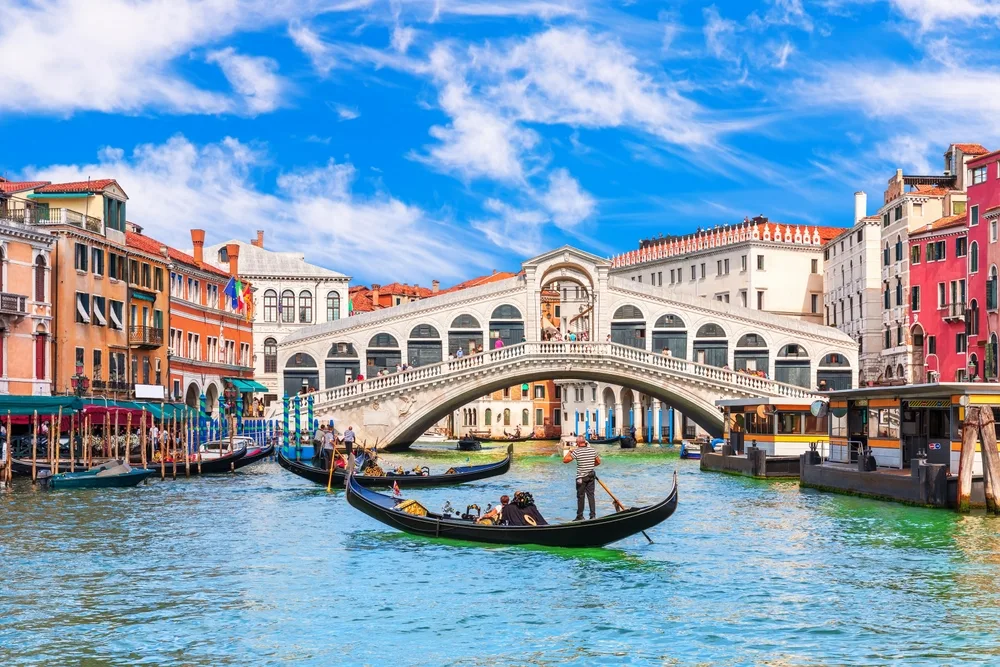 View of the Rialto Bridge over the Grand Canal in Venice during a beautiful day with gondolas on the water for a list of the best cities to visit in Italy