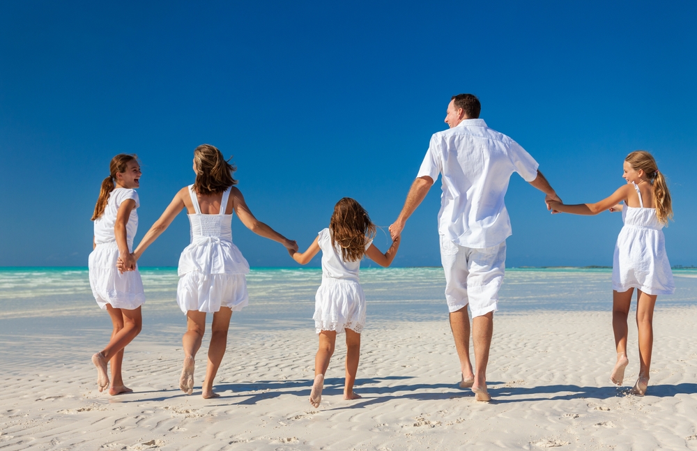 A family of five, all wearing white on the beach, holding each other's hands while running on the shore. 