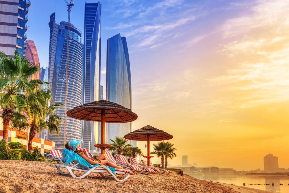 Woman in a swim coverup lying on a beach chair on the sand with a hazy coastline in the background and a yellow sky overhead for a guide titled Is Dubai Safe to Visit