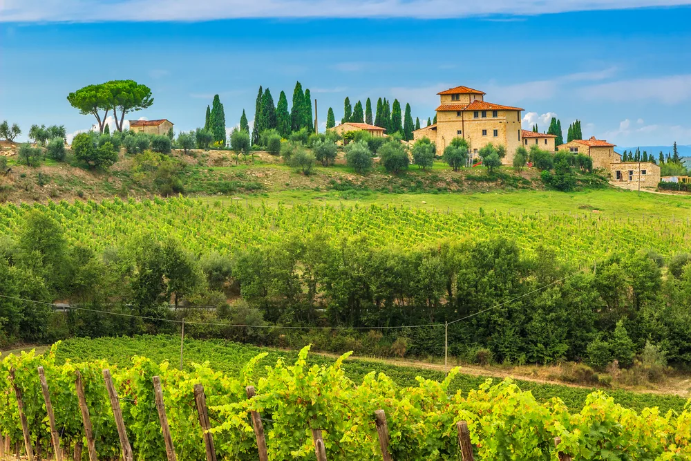 Beautiful traditional home on a hill in the Tuscany Chianti region with vineyard rows in the foreground and lush greenery for an FAQ section covering the best cities to visit around Italy