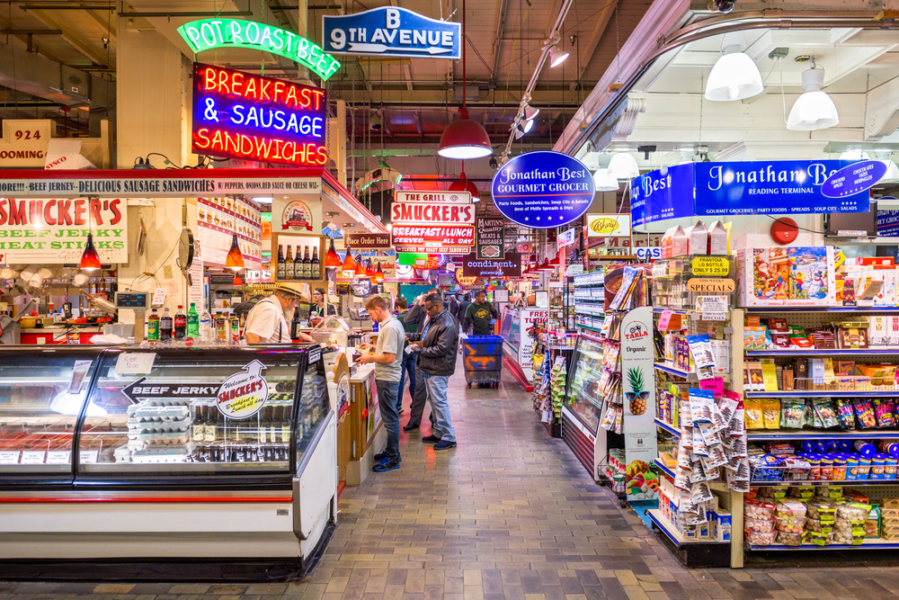 People crowding the Reading Terminal Market in the middle of the day below neon signs and shelves of stuff for a guide titled Is Philadelphia Safe to Visit