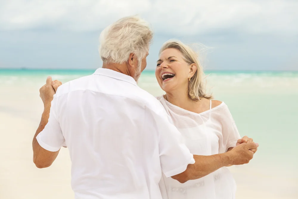 Two old couple wearing white clothes, dancing on the beach.