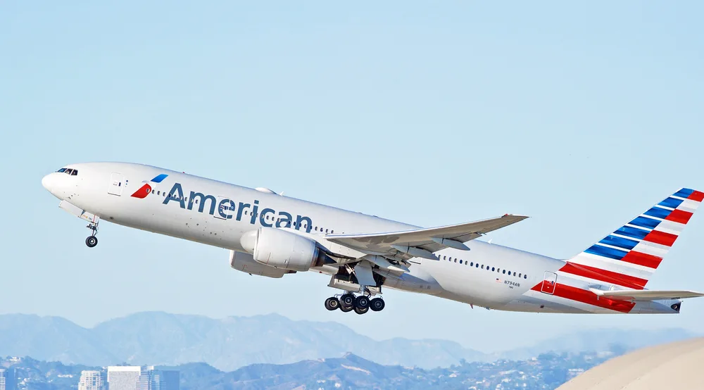 A plane that just took off with a label American and in background are mountains and buildings. 