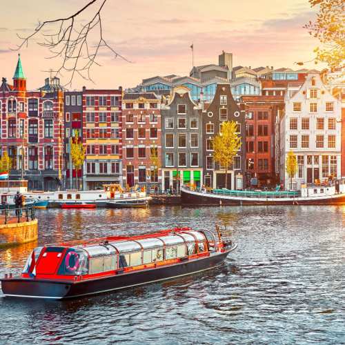 a ferry cruising along a wide river at the center of the city where on its banks are old buildings during a sunset if the best time to visit Netherlands.