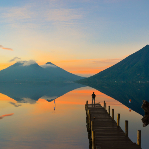 a person standing on a wooden dock while looking at the tall mountains reflected on the lake during a sweet sunset on one of the best time to visit Guatemala.