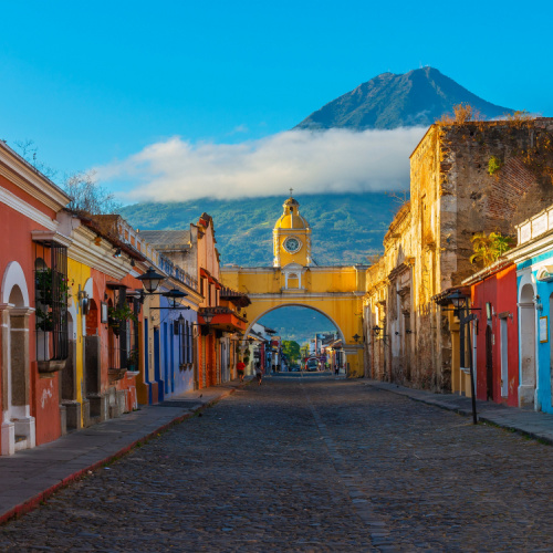 a street in a country town where an arch with a cross can be seen, and in a far distance is a volcano with a cloudy peak during the best time to visit Antigua.
