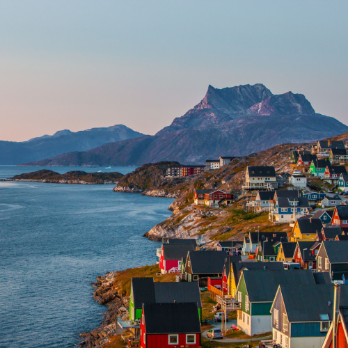 a coastal town where the houses seems like in the same design and facing the sea, and on the distance are tall mountains, during a sunset of the best time to visit Greenland.