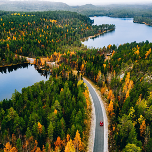 a calm scenery where a car is drivin on a road surrounded by trees and passing by a lake, during the best time to visit Finland.