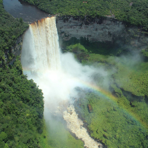 top view of a large waterfalls surrounded by a vast forest, and a rainbow can be seen at the foot of the waterfalls, photographed during one of the best time to visit Guyana.