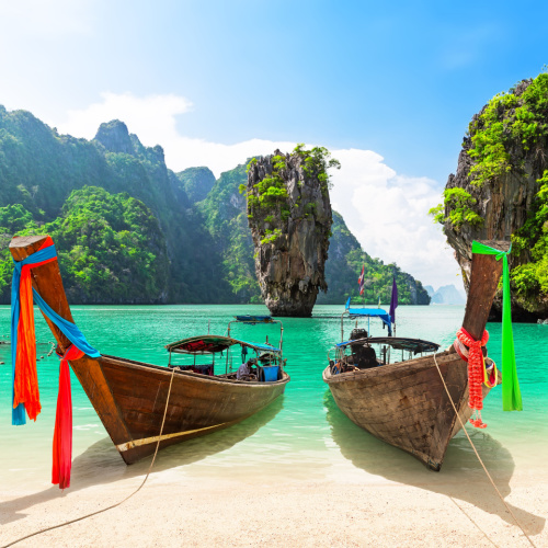 two traditional boats docked on the shore of a beach where a large rock can be seen standing above the emerald waters, seen during the best time to visit Thailand.