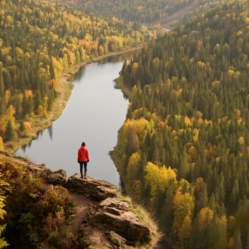 a view from a peak of a mountain where a woman wearing red is standing on a rock with an overlooking view of a river and a forest during the best time to visit Russia.