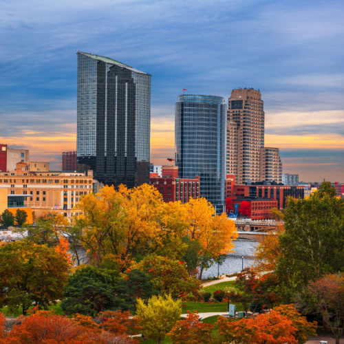 a view from the park where the leaves are in the tint of orange during the autumn season of the best time to visit Michigan, where visit in background are buildings of the city during a sunset.