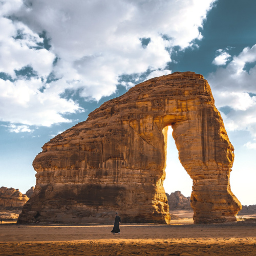 a person standing in front of a huge rock formation in a desert during the best time to visit Saudi Arabia.