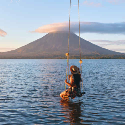 a woman on a swing where her legs are touching the seawater, staring at the tall mountain with cloudy peak during a sunset on one of the best time to visit Nicaragua.