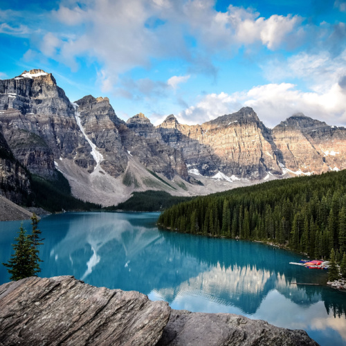 a view of a lake with calm and blue water where on one side is a forest and on the other side are mountains, seen during the best time to visit Canada.