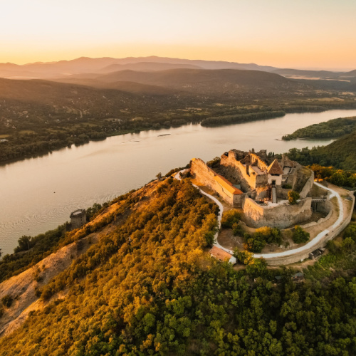 a castle built on top of a hill surrounded by trees, and at the bottom is a wide river, photographed on a summer of the best time to visit Hungary.