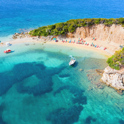 the end of an island where several boats can be seen on it clear waters, and several sunbeds on its white shore, during a hot afternoon of the best time to visit Albania.