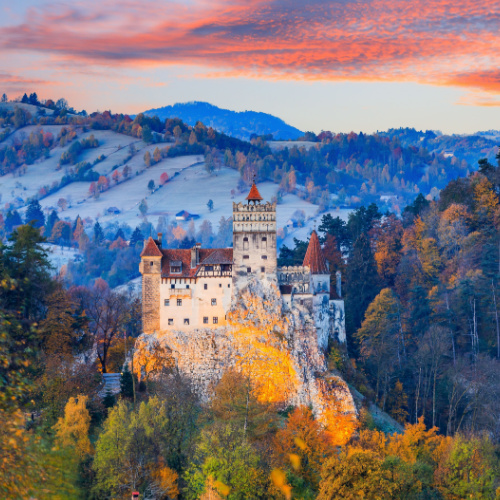 a castle on top of a hill surrounded by forests and in background are mountains during a sunset of the best time to visit Romania.