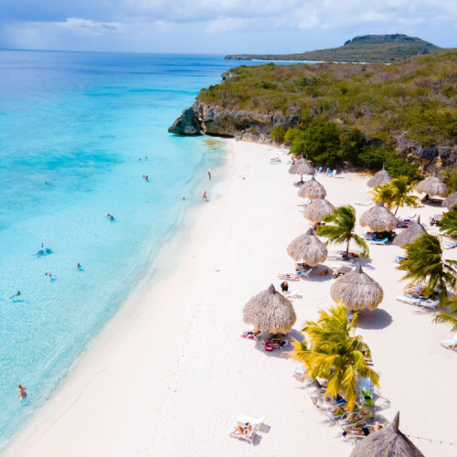 aerial view of a beach with white sand and clear waters, and native structures can be seen on the beach during a hot afternoon of the best time to visit Curacao.