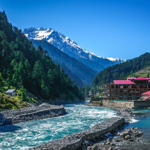 a gushing river where its riverbanks are filled with trees, and several structures, and in background is an icy mountain during the best time to visit Pakistan.