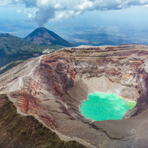the crater of a volcano where a greenish liquid can be seen, and in background is another smoking volcano, photographed during one of the best time to visit El Salvador.