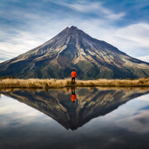 a person standing facing a volcano, perfectly refelected on the still lake, during the best time to visit New Zealand.