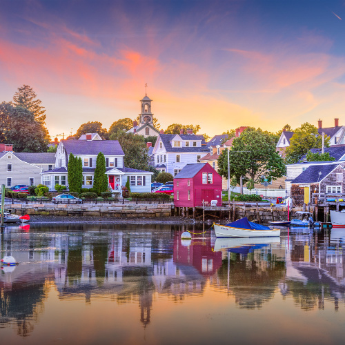 a town in the coastal area where the houses are in light colors and boats can be seen docked on a pier, photographed during the best time to visit New Hampshire in a sunset.