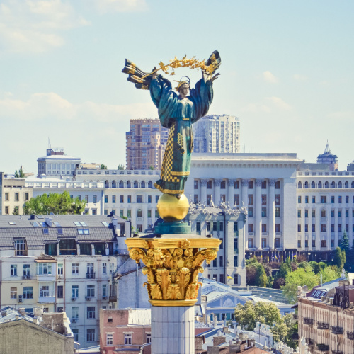 a statue on top of a column where the figure looks like a women raising a flower, and in background are city buikdings during the best time to visit Ukraine.