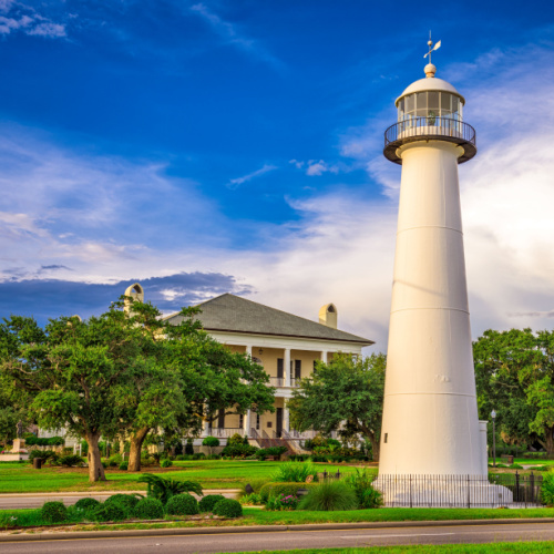 a lighthouse surrounded by a park where the trees and grass are green, where a large house can be seen behind the trees, photographed during the best time to visit Mississippi.
