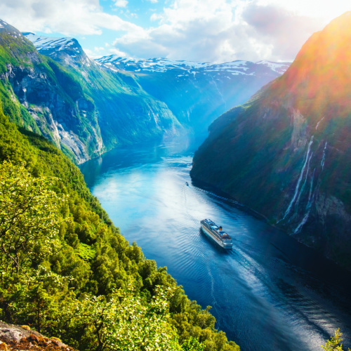 a cruise ship passing by a river in betwwen mountain ranges with a small waterfall on the side during a cloudy afternoon of the best time to visit Scandinavia.