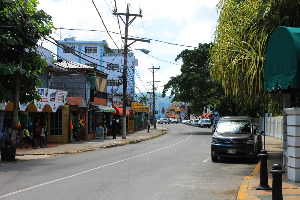 For a piece on the best and worst times to visit Montego Bay, a photo of an empty road in the town pictured with a few cars parked on the side of the road, gloomy skies overhead, and little shops with their doors open